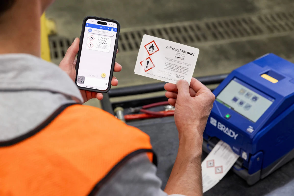 A printed chemical hazard label beside the Brady i4311 and a smartphone